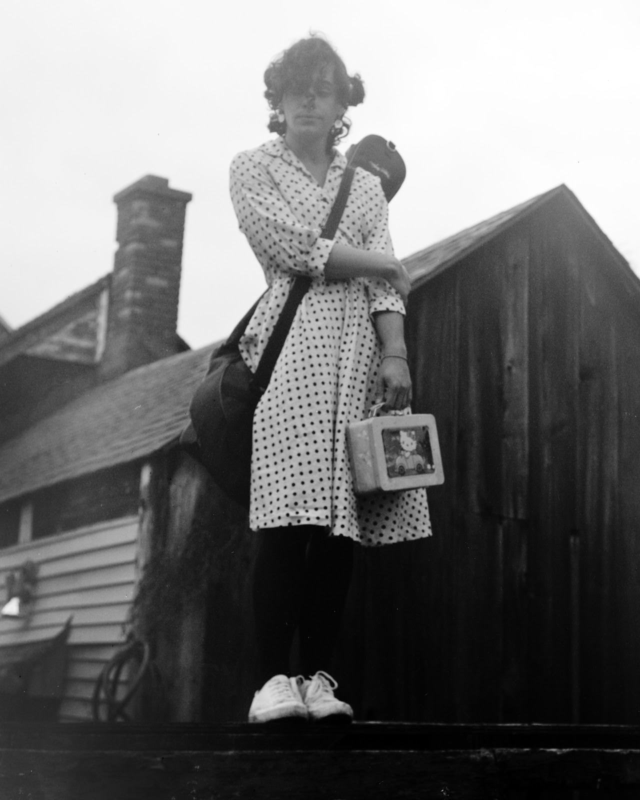 Alex Merkel's headshot. Black and white. He stands balancing on a fencepost looking down with one arm crossed over his chest holding the other. In the other hand, he holds a small "Hello Kitty" tin lunchbox. A black guitar case is strapped across his chest, with the guitar resting on Alex's back. His facial expression is peaceful.