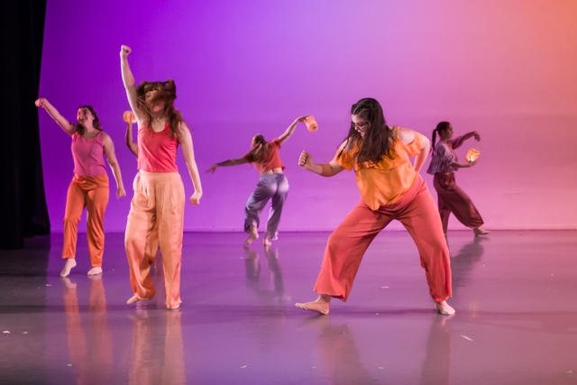 Five smiling dancers spread across a stage lit in purple and pink, wearing pink, orange, and lavender costumes, with back dancers holding lanterns