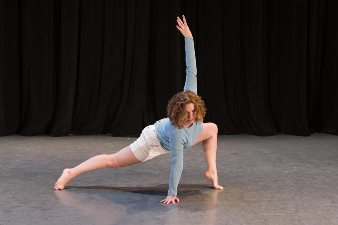 Hannah posing in a runner's lunge position on the floor, with one arm extended above her head and the other supporting her weight on the floor. She looks fiercely towards the camera, and her curly hair covers part of her face.