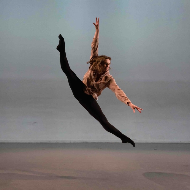 Hannah executing a tilt jump with pointed toes and arms positioned in an L-shape, looking down over her left arm