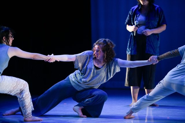 Hannah in a deep side lunge holding hands with two dancers on either side while looking into the eyes of the dancer on her right, with another dancer behind her holding a microphone