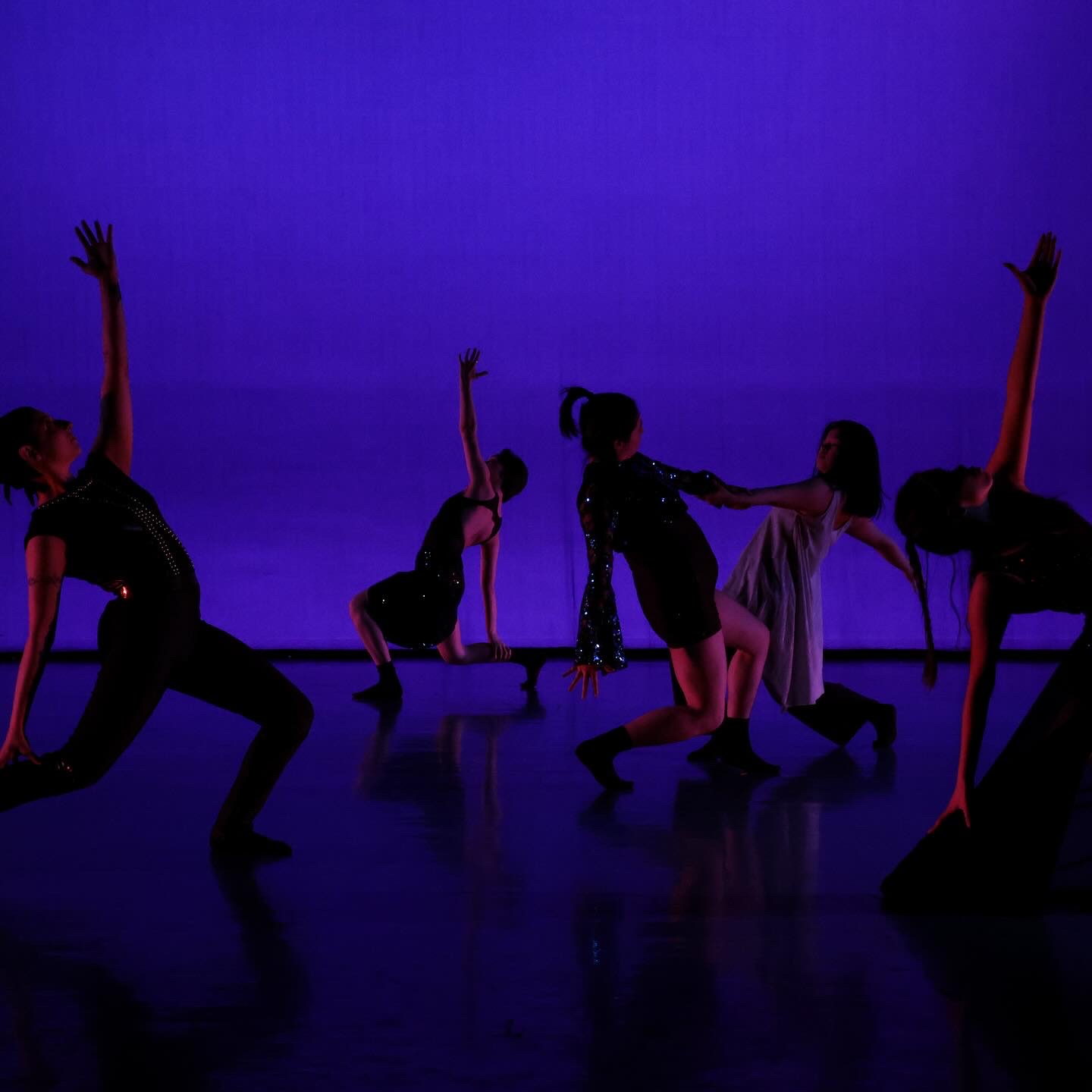 Five dancers on a dimly-lit stage in shimmering costumes, three in lunges with arms perpendicular to the floor, two in center holding hands in counter-balance