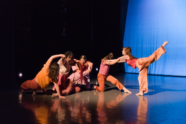 Six dancers in pink and orange engaging in contact improvisation, four kneeling and two standing, with one dancer on the right lifting their leg in attitude to the back
