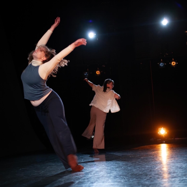 Hannah falling gracefully to her right side with arms extended overhead and back arched, eyes closed and face toward the light, with another dancer balancing on one leg behind her
