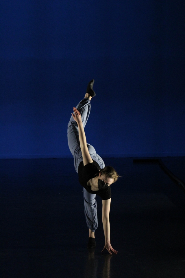 Hannah performing an illusion step with one hand on floor, right leg in attitude above her head, wearing black mask, black leotard, light blue pants, and black socks
