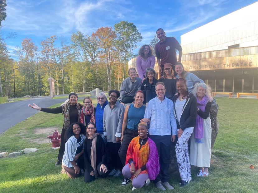 A diverse group of seventeen adults pose in lines for a photo. The group is outdoors in front of the Doris Duke Theater at Jacob's Pillow, wearing a variety of colorful outfits, and smiling.