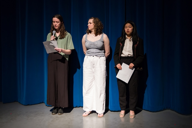 Three people dressed nicely stand in front of a blue curtain, looking out into the crowd (offscreen).