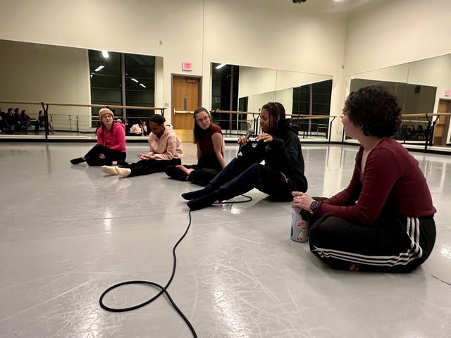 Five MHC students sit on the floor in a semicircle, looking at one another in conversation. One of them is holding a microphone and speaking.