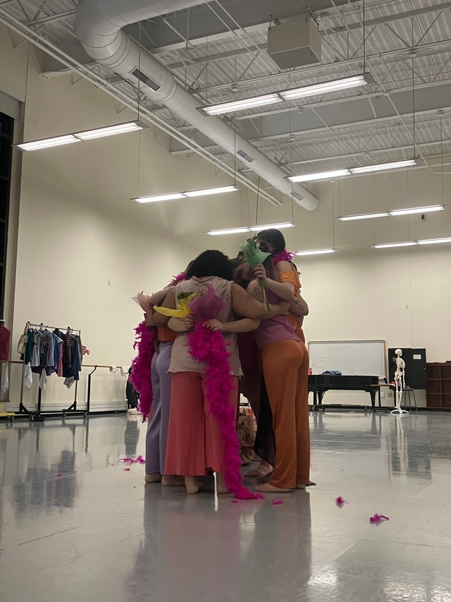 A group of dancers huddle in a circle with their arms around another, holding bright pink feather boas and bouquets of flowers in their hands.