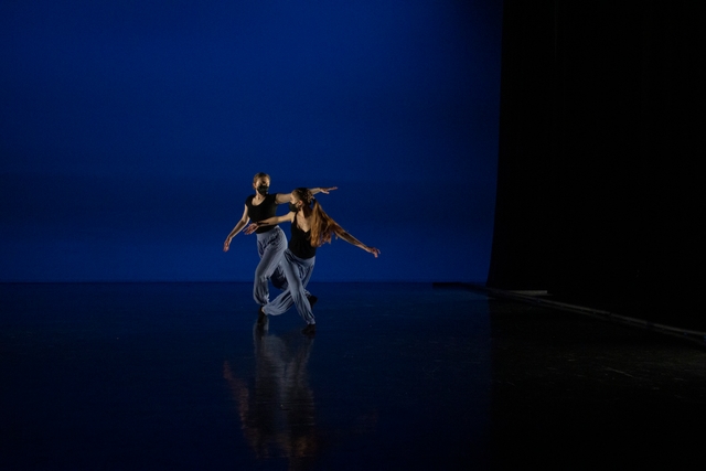 Two dancers stand next to each other, with their legs in lunged positions. They look at each other with their arms outstretched to either side.