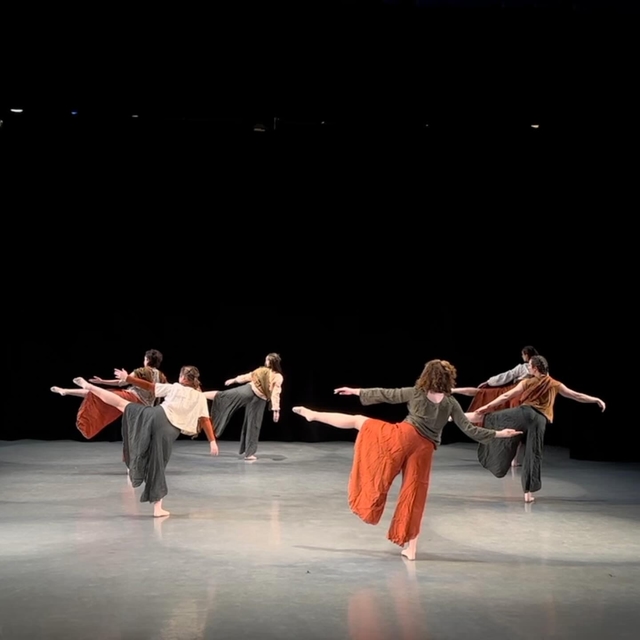 Six dancers stand spread across a stage, facing away from the camera with their left legs lifted to their sides in an "attitude" position, with their toes forward, and their arms outstretched parallel to their legs in the air.