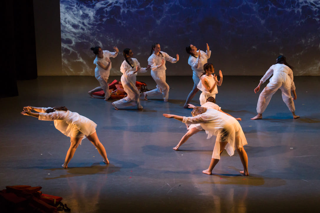A group of dancers is spread across a dimly lit stage with an image of ocean waves projected in the background. The dancers in the back (including Hannah) hold their hands in front of their faces. With their palms facing outward. The dancers towards the front of the shot are facing away from the camera with their upper bodies pitched forward and arms stretched out.