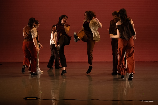 Hannah jumps in the air with her leg kicked out in front of her, smiling at another dancer doing the same thing beside her. Other dancers stand around them forming two vertical lines on either side, also smiling. Metal taps are visible on the bottoms of the centered dancers' shoes.