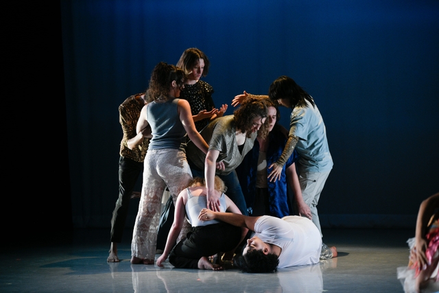 A group of eight dancers are standing and sitting on the ground very close to one another, holding their bodies in various positions wrapped around one another. Hannah is at the center of the mass, standing up and leaning forward while looking down.