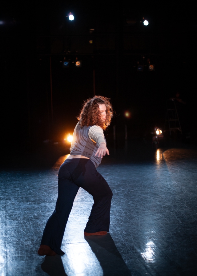 Hannah stands with one leg crossed in front of the other and bent. Her arm is stretched toward the camera, and she gazes softly beyond it, toward the right of the screen.