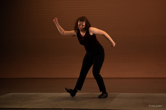 Hannah stands centered in the frame, with her legs bent slightly and her arms bent at 90 degree angles, flipped in opposite directions. She smirks and looks at the floor.