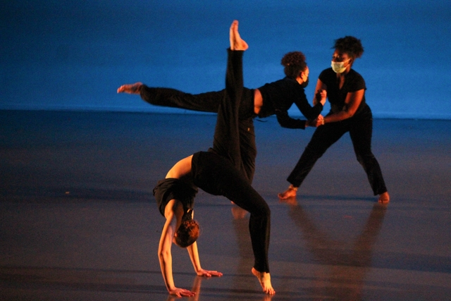 Hannah (front) arches her back with her hands touching the floor. One leg is kicked up vertically, and the other is just leaving the floor. Two dancers in the background hold hands facing each other while one has her leg outstretched behind her.