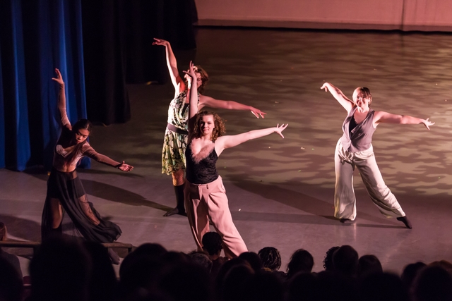 Four dancers stand close to the audience, facing them. They each have one leg straightened and extended on the floor with their toes pointed to the side. Their arms form an "L" shape above their heads, and they smile while looking out.